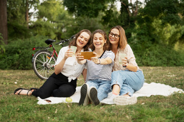 Fototapeta premium Three beautiful happy young women close friends relax in a green park on a blanket. Have fun and chat on the green grass. Taking a selfie on a smartphone