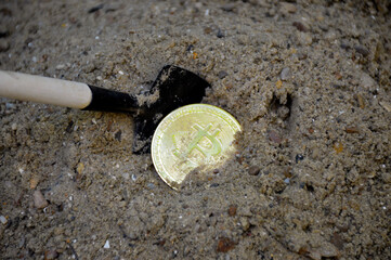 A gold bitcoin coin is half buried in the sand with a shovel next to it