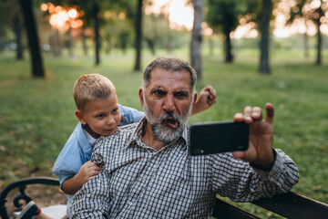 boy with his grandfather sitting on the bench and using mobile phone in the city park