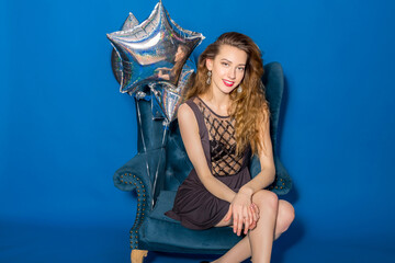 Obraz na płótnie Canvas pretty cool young hipster beautiful woman, evening dress, trend fashion style, blue background, sitting in chair, silver balloons, smiling, happy, celebrating birthday