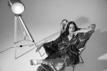 Fototapeta na wymiar Young fashion female model with long straight brunette haie and makeup with pink eye shadows wearing silver kimono with trousers and white clog shoes sitting with crossed legs in stylish armchair