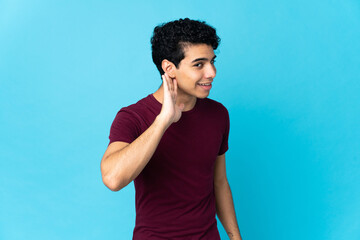 Young Venezuelan man isolated on blue background listening to something by putting hand on the ear