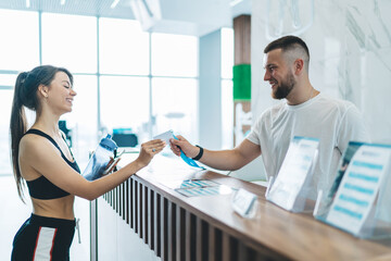Cheerful caucasian male receptionists talking with slim client giving e-key for access to locker room and gym, smiling girl entering training center taking card from happy hospitable man worker