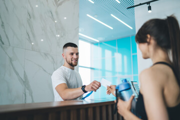 Handsome caucasian man receptionist in gym greeting female client giving key to access for training, happy male employee communicating with 20s woman taking card for opening locker room - Powered by Adobe