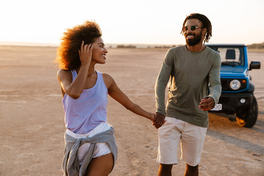 Image of joyful african american couple holding hands together and walking on desert