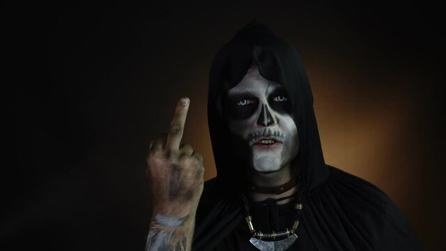 Sinister man with skull makeup making faces and showing middle finger. Bad manner gesture. Halloween