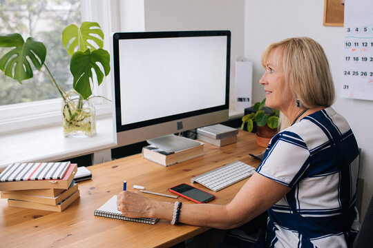 Blonde Mature Woman By Computer. Working From Home Office. Computer With Blank Empty Screen For Copy Space And Information. A Businesswoman From Behind Shoulder View