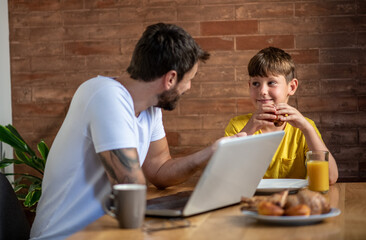 Father and son spend time together in the kitchen during the morning, father turns on the computer to check online post while son has breakfast, they talk