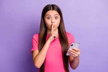 Photo portrait of small sweet little girl with long brunette hair keeping secret holding cellphone touching lips with finger isolated on purple color background