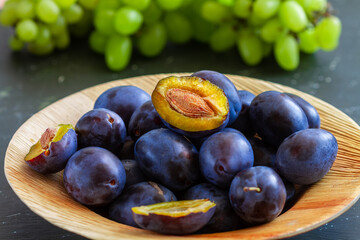 Ripe and fresh black plums lie in a wooden plate and stand on a black table.