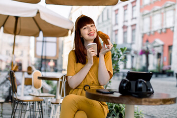 Beautiful young Caucasian red haired woman, sitting in outdoor city cafe, holding paper coffee cup and fresh tasty croissant, while having her lunch time. Business lady outdoor in city cafe