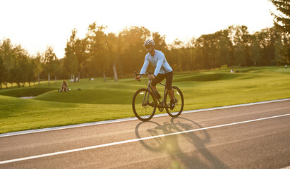 Side view of a young athletic man in sportswear cycling in city park at sunset, riding mountain road bike