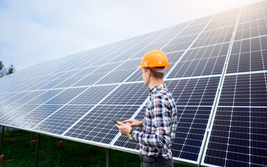 Engineer in helmet with tablet in hands standing near solar panels on green plantation. Concept...