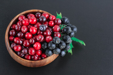 Cranberries and chokeberry in wooden bowl on black background. Nature, autumn, crop, food, berry concept.