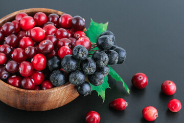 Cranberries and chokeberry in wooden bowl on black background. Nature, autumn, crop, food, berry concept.