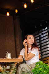 Young caucasian woman sits on street area of cafe, at wooden table with cup, with yellow bulbs and flowers in summer