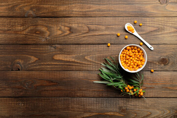 Sea buckthorn berries in bowl, spoon and plant leaves on wooden table. Flat lay composition, top view.