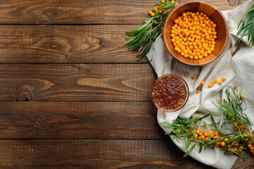 Sea buckthorn berries in wooden bowl, sea buckthorn jam and plant leaves on wooden table. Flat lay composition, top view.