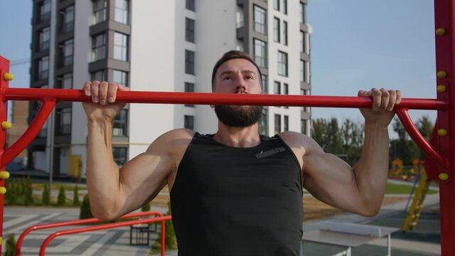 A young man of athletic build pulls up on a red horizontal bar. Against the background of the sports ground.