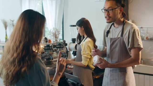 Young Asia male barista waiter taking order from customer standing behind bar counter while talking with customer making note on takeaway coffee cup at cafe restaurant. Owner small business concept.