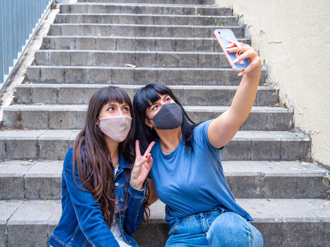 Two caucasian young women sitting outside taking a selfie with cellphone and wearing face masks during COVID. Friendship, millennial and coronavirus concept. Girl doing peace sign for a photo.