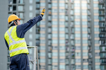 Asian maintenance worker man with safety helmet and reflective suit standing on aluminium step ladder at construction site. Civil engineering, Architecture builder and building service concepts