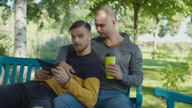 Positive handsome male homosexual drinking hot drink from travel mug, hugging his beloved partner busy networking with tablet pc and chatting while joyful same-sex couple resting on park bench.