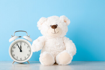 White alarm clock and smiling teddy bear on wooden table at light blue wall background. Pastel color. Time concept. Closeup. Front view.