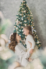 Pretty little girl and beautiful mother in white sweaters hug near christmas tree and make hairstyle together