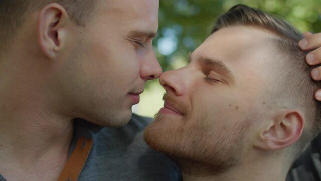 Portrait of affectionate handsome gay couple expressing love, happiness and warm relationship by rubbing their noses and gently kissing during romantic date in public park on sunny summer day.