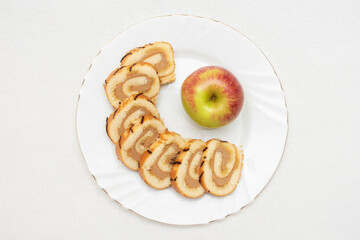 Sponge roll cut into pieces and red apple on a white plate, top view, copy space