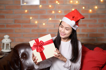 Young smiling woman wearing red Santa Claus hat showing a gift box on Christmas day, holiday concept.