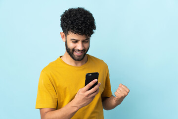 Young Moroccan man isolated on blue background using mobile phone and doing victory gesture