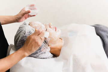 Obraz na płótnie Canvas Beautician hands making anti-age procedures applying foam cleansing mask for mid-aged female client at beauty clinic.