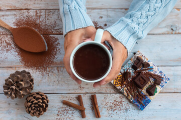 Directely above view of two female human hands  holding a chocolate cup, creamy and thick. Wooden table as background with pieces of dark chocolate, spoon and pine cones