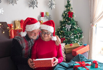 Fototapeta na wymiar Cheerful couple of senior people sitting on the sofa with Santa Claus caps opening a present with Christmas tree and gifts for the family in the background. Love and family concept