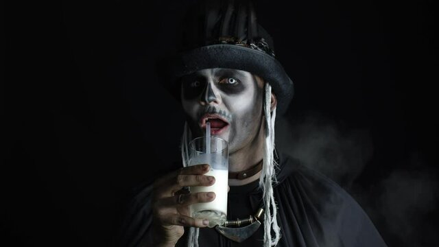 Scary guy in carnival costume of Halloween skeleton looking at camera, drinks milk from a glass