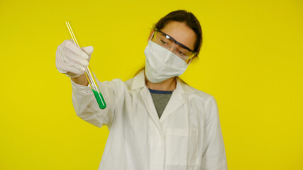 Young doctor in medical mask goggles latex gloves looks tests in tube. Woman in white coat on yellow background holds hand tube with green liquid