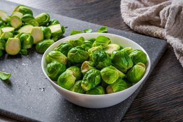 Fresh raw brussels sprouts in a white bowl on a dark cutting board with a towel at the background and wooden table