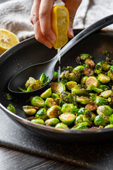A hand squeezes lemon juice over roasted / fried brussels sprouts in a black frying pan that sits on a cutting board with a solid spoon for serving. Half a lemon and towel in the background