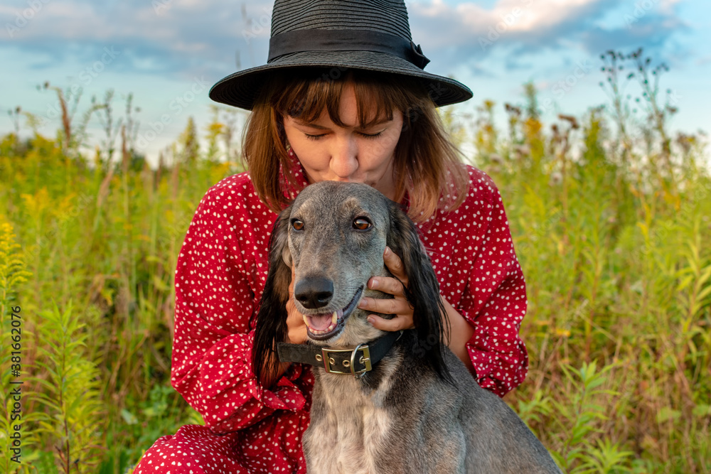 Wall mural The girl with her pet are walking in the park. A young beautiful woman in a red dress and hat kisses her dog on the forehead