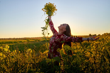 Beautiful woman in red dress holding bouquet of yellow flowers and enjoying summer sunny day in green field with green grass in sunset with nice orange sun