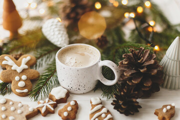 Cozy winter mood. Coffee with gingerbread cookies, pine cones and warm lights on white wooden table