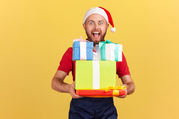 Extremely happy courier in blue uniform and red santa claus hat holding and showing christmas gifts looking at camera with toothy smile. Indoor studio shot isolated on yellow background
