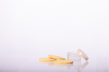 cosmetic cream jar and Three Beige round sponge pads for face make-up cleaning reflected in glass surface isolated on white background.