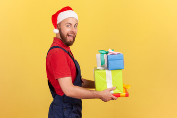 Side view extremely happy delivery man in blue uniform and red santa claus hat holding present boxes looking at camera with big eyes and open mouth. Indoor studio shot isolated on yellow background