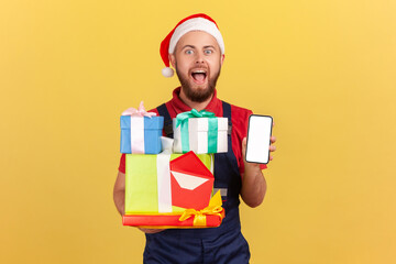 Excited surprised courier in uniform and santa claus hat holding and showing gift boxes and smartphone with empty display, order application. Indoor studio shot isolated on yellow background