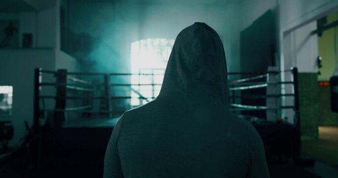 Cinematic slow motion back view shot of a professional young man wearing grey hoodie is entering in a boxing ring for starting his workout.