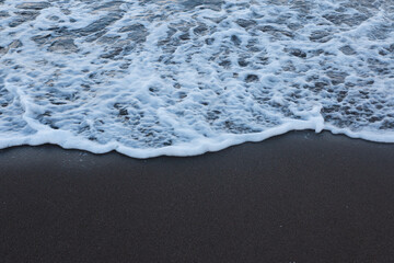Wave of the sea calmly reaching the shore of the beach with black sand.