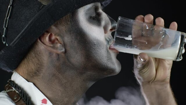 Scary guy in carnival costume of Halloween skeleton looking at camera, drinks milk from a glass